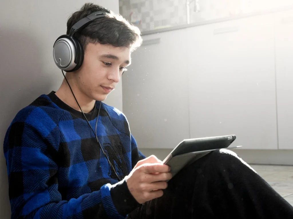 Teen with headphones, sitting on the floor and focused on a tablet. Sunlight softly illuminates a modern kitchen in the background